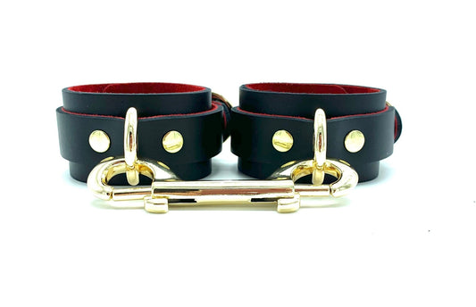Front view of black leather handcuffs with red suede lining, featuring a gold connector that highlights their luxurious design, ideal for BDSM enthusiasts looking for stylish and versatile restraints, crafted in the USA.