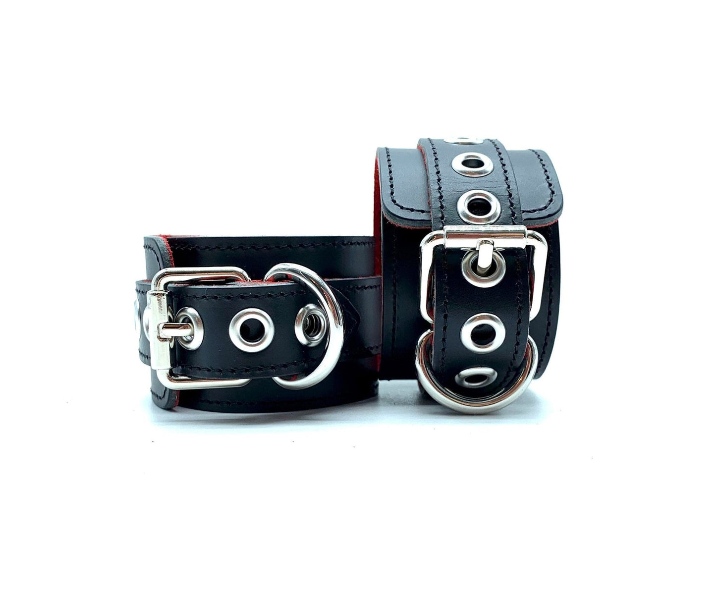 Pair of Italian BDSM bondage cuffs in Scarlet design, featuring one cuff lying flat and the other positioned upright, highlighting the shimmering silver buckles against the contrast of black leather and red suede, ensuring durability and elegance in bondage encounters.