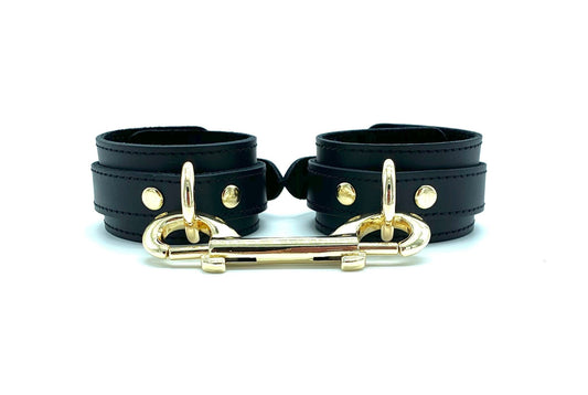 Front view of Tango black leather cuffs with black suede lining and black stitching, complemented by gold hardware and a gold connector, showcasing a luxurious and cohesive design ideal for sophisticated BDSM play.