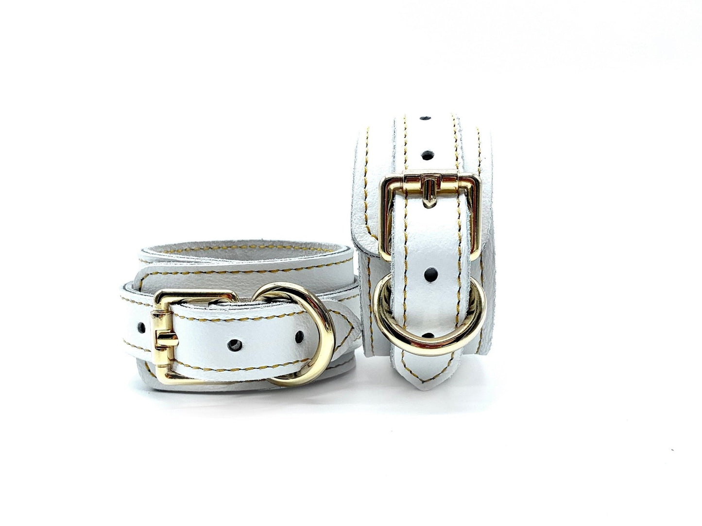 Pair of Italian white leather and suede BDSM bondage cuffs made in the USA, featuring one cuff lying flat and the other positioned upright, highlighting the gleaming gold hardware, offering both elegance and durability for captivating bondage experiences.