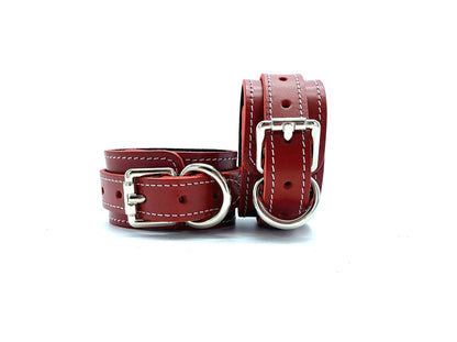 Two red leather handcuffs displayed side by side from the Tango Red BDSM Kit, one standing upright and the other lying flat. Both cuffs feature silver buckles and are detailed with black suede and white stitching, showcasing their high-quality construction and elegant design