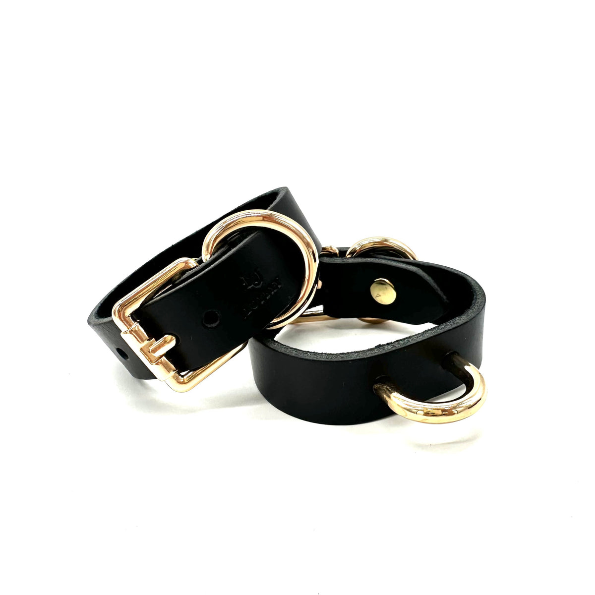 A pair of black leather wrist cuffs showcasing gold buckle closure and centered D ring.