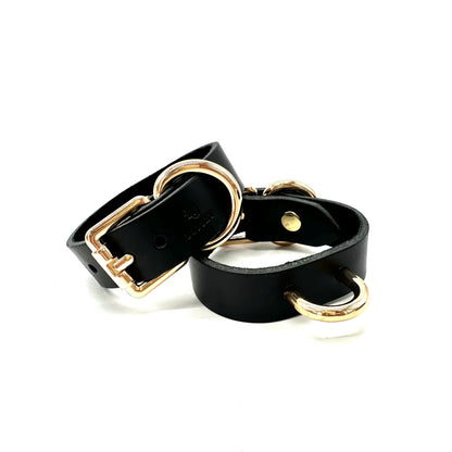 A pair of black leather wrist cuffs showcasing gold buckle closure and centered D ring.