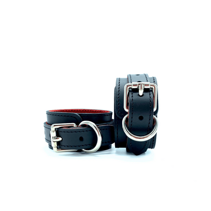 A pair of black leather handcuffs with red suede lining, detailed stitching and silver hardware on white background.