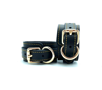 Close up of black leather handcuffs featuring detailed gold stitching and elegant gold buckle.