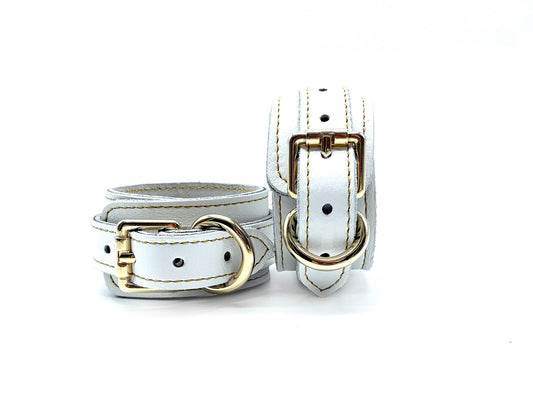 Pair of white Italian leather bondage cuffs with gold hardware, one flat and one upright showing adjustment holes