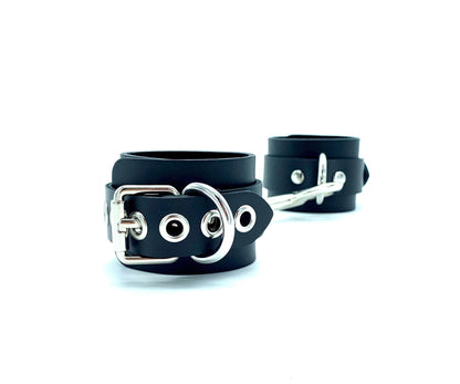 Pair of Italian 2" BDSM bondage cuffs made in the USA, featuring black leather and black suede cuffs connected sideways, with one lying flat and the other positioned upright, revealing the shimmering silver hardware, ensuring both style and functionality for intimate bondage play.