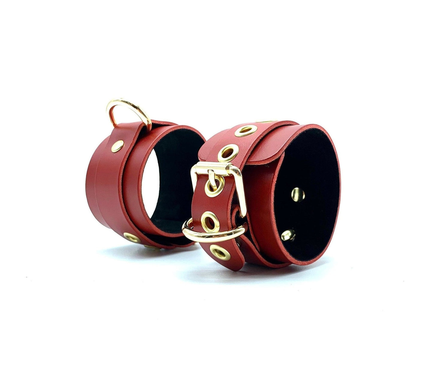 Red leather cuffs with black suede lining, standing upright next to each other, showcasing their sleek gold hardware, designed for visual appeal and effective performance in bondage settings.