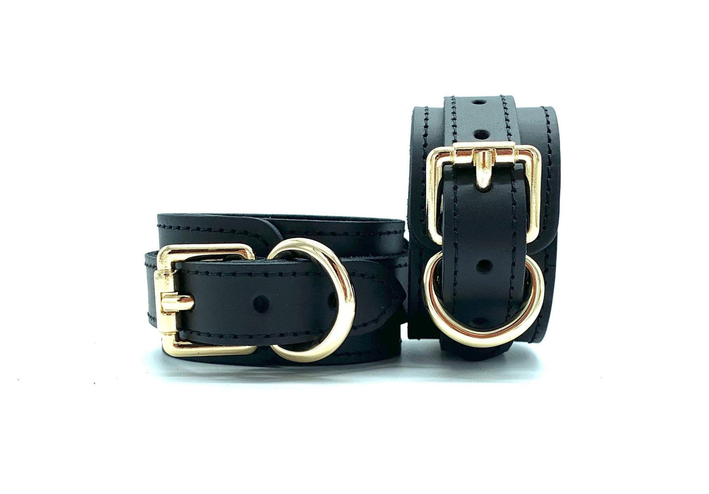 Front view of Tango black leather cuffs, one standing upright and the other lying flat, both featuring striking gold hardware, illustrating the versatility and elegant construction suitable for diverse BDSM activities.